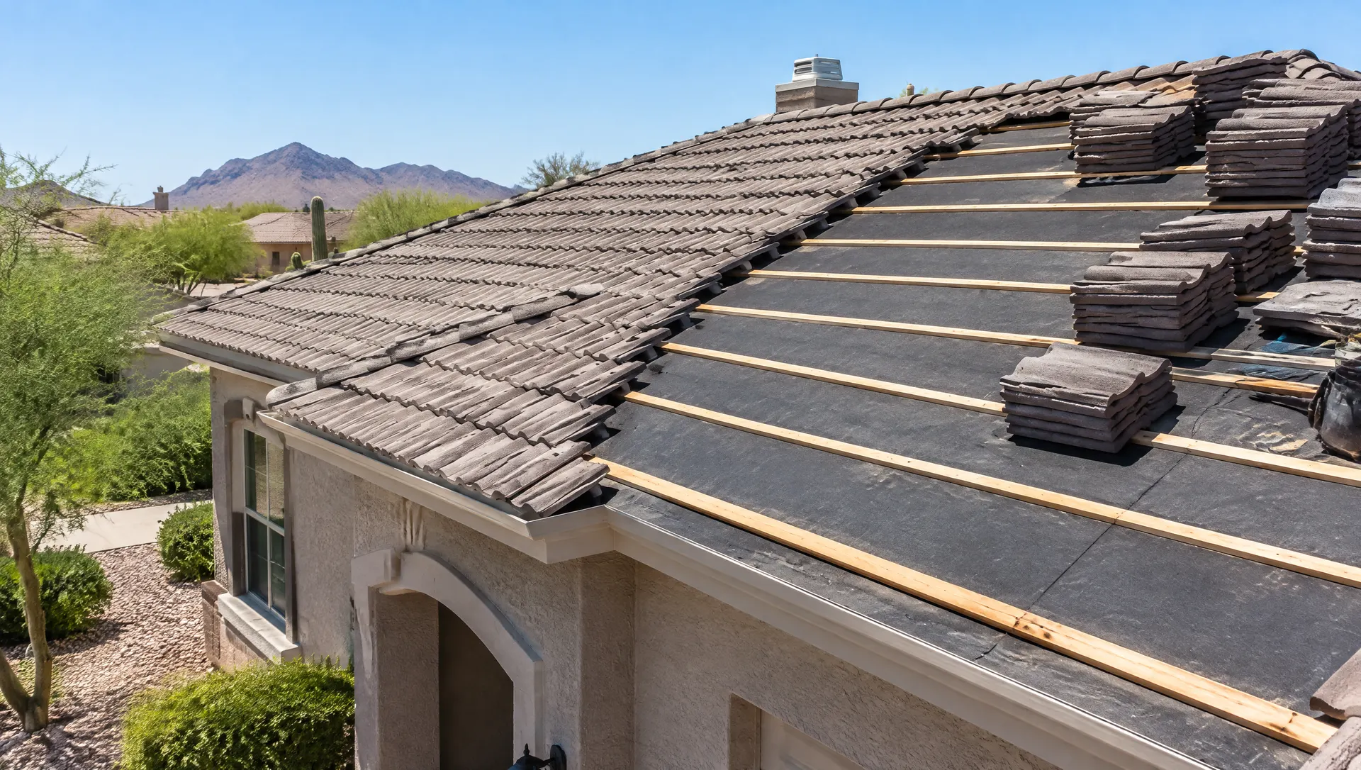 Roofers' work in progress on a concrete tile roof: one section of tiles lifted and neatly stacked on the roof, new dark underlayment and wood battens exposed, the rest of the roof still tiled, bright Arizona sun